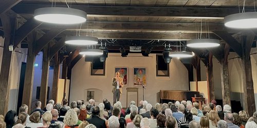 In einem Saal sitzen viele Menschen im Publikum und schauen Richtung Bühne, auf der eine Person im Anzug steht.